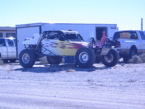 1990 Ford Bronco - white off-road race buggy with flame graphics parked between an enclosed trailer and white pickup trucks in the desert.