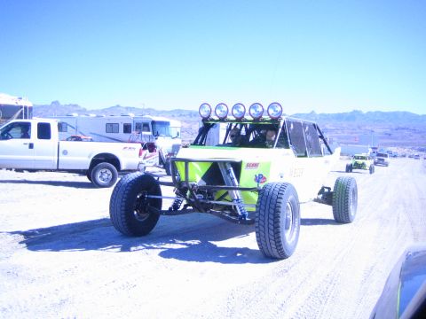 1990 Ford Bronco - green and black tube-frame off-road race buggy with five roof-mounted lights drives on a dusty desert road past trucks and an RV.