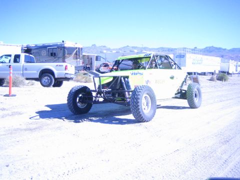 1990 Ford Bronco - green and white tube-chassis off-road race buggy sits on packed desert dirt beside a white pickup truck and motorhome in a pit area.