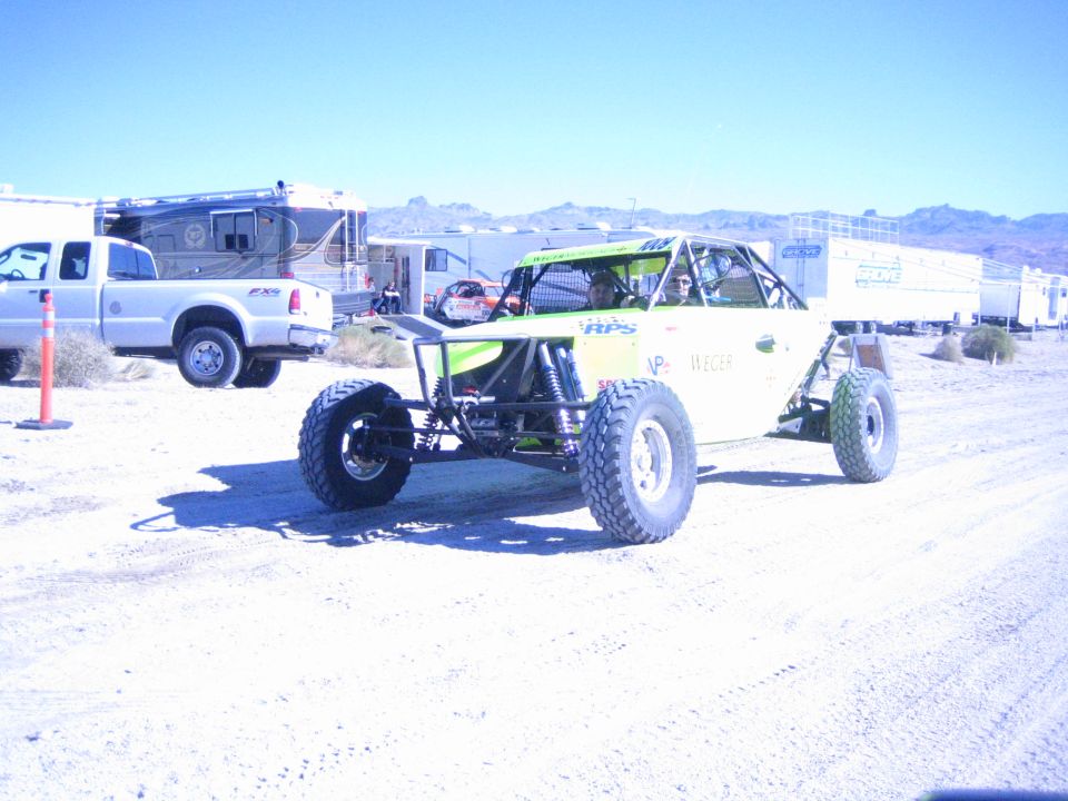 Green and white off-road buggy with long-travel suspension on desert dirt near a white pickup and RV