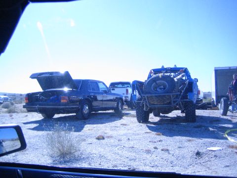 1990 Ford Bronco - Shot through a windshield, a black sedan with its trunk open sits beside a blue off-road buggy and enclosed trailer in the desert.