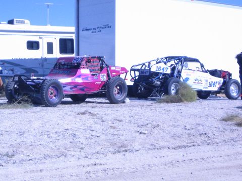1990 Ford Bronco - pink flame-painted off-road buggy number 902 and a blue tube-chassis buggy number 1647 sit on dirt beside a racing trailer.