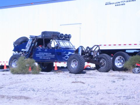 1990 Ford Bronco - blue flame-painted off-road race buggy lettered Kevin McMullen Fabrication / Transaxles sits on dirt next to a flatbed trailer.
