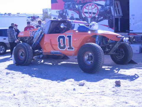 1990 Ford Bronco - orange off-road race buggy numbered 01 in a General Lee style sits in a desert pit area as people gather around it.