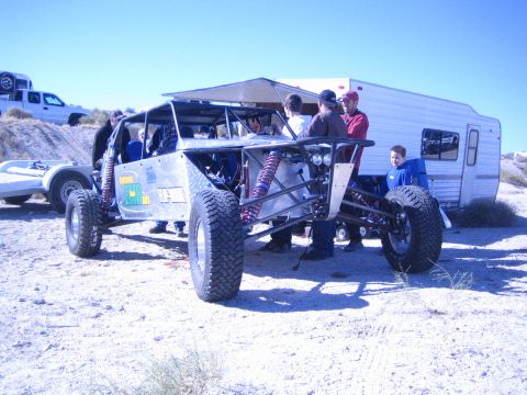 1990 Ford Bronco - tube-chassis off-road buggy with an awning shade sits beside a travel trailer in the desert as a group of people stand around it.