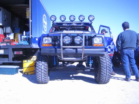 1990 Ford Bronco - front of a lifted blue Jeep Cherokee off-road build, with roof light bar and large tires, sits in a pit beside a tool bench and trailer.