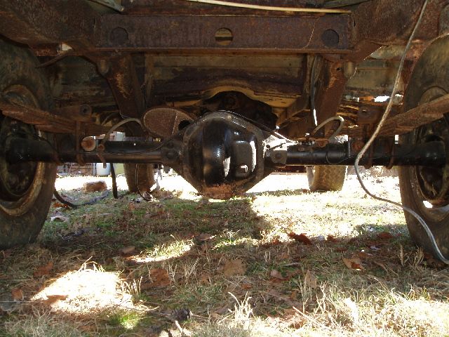 Rear axle and differential housing under a rusty vehicle chassis, viewed from behind over grass.