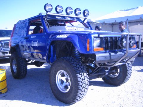 1990 Ford Bronco - lifted blue Jeep Cherokee with roof-mounted light bar, custom graphics and large off-road tires is parked on gravel under event tents.