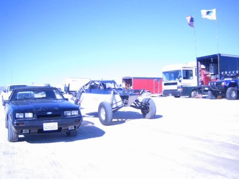 1990 Ford Bronco - black 1980s Ford Mustang notchback is parked beside an off-road buggy on a dry desert lakebed with trailers and flags behind.