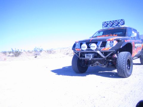 1990 Ford Bronco - black Nissan off-road race truck with PIAA lights, light bar and front winch bumper sits on open desert flats.