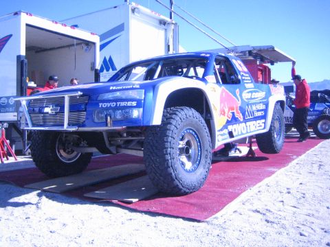 1990 Ford Bronco - blue Chevrolet-bodied trophy truck with Red Bull and Toyo Tires graphics is displayed on a red carpet beside an enclosed trailer.