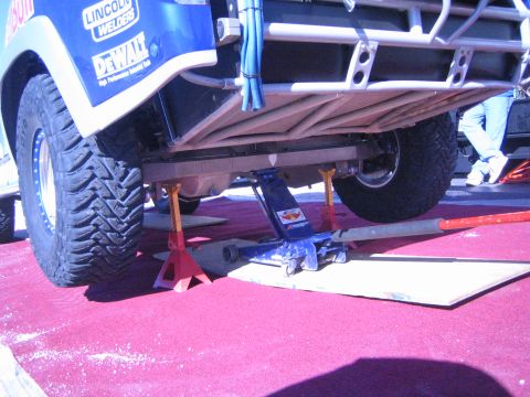 1990 Ford Bronco - underside of a blue race truck is shown raised on a floor jack and jack stand over a red carpet, exposing tires and chassis.