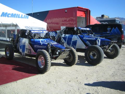 1990 Ford Bronco - Two blue and white tube-chassis off-road race buggies with BFGoodrich graphics sit parked under McMillin Racing tents on dirt.