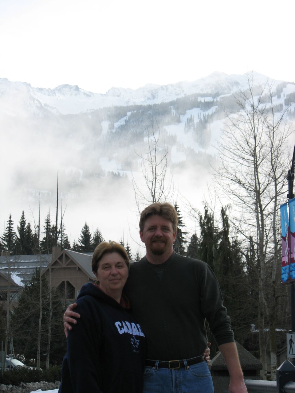 Man and woman posing outdoors in front of fog-shrouded snowy mountain and lodge