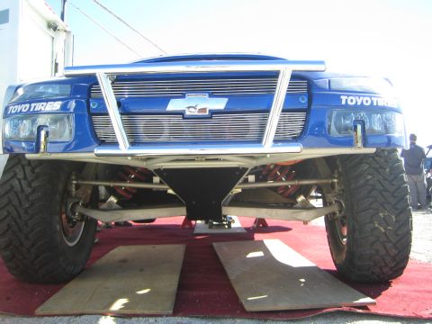 1990 Ford Bronco - Front view of a lifted blue Chevrolet-bodied race truck on Toyo Tires, raised on ramps over red carpet showing the chassis underneath.