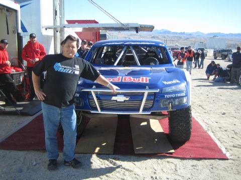 1990 Ford Bronco - man stands beside a blue Chevrolet-bodied Red Bull trophy truck displayed on a red carpet in a desert pit area as crowds mill about.