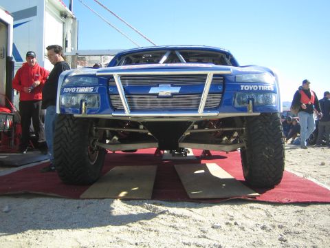 1990 Ford Bronco - low front view of a blue Chevrolet-bodied Toyo Tires race truck raised on ramps over red carpet as people stand around it.