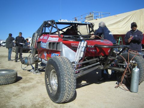 1990 Ford Bronco - red tube-chassis off-road race truck sits in a desert pit with crew working beside it under a clear blue sky.