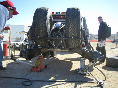 1990 Ford Bronco - race truck is lifted on floor jacks in a desert pit, exposing its underside and chassis as a man with a backpack watches.