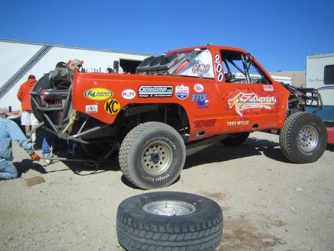 1990 Ford Bronco - orange off-road race truck marked 800 with AZ Land Racing decals sits in a dusty pit near trailers.