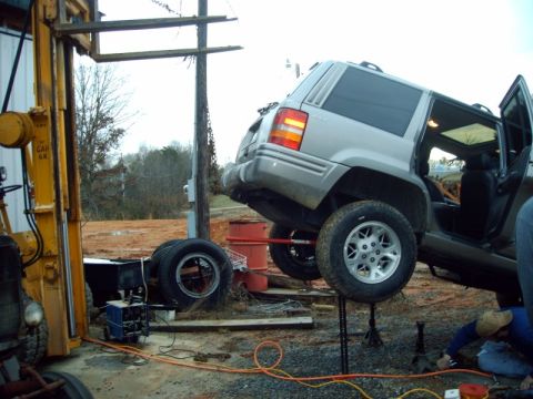 2005 Ford Excursion - A Jeep Grand Cherokee is being worked on, lifted by jacks and a forklift in an outdoor repair area.