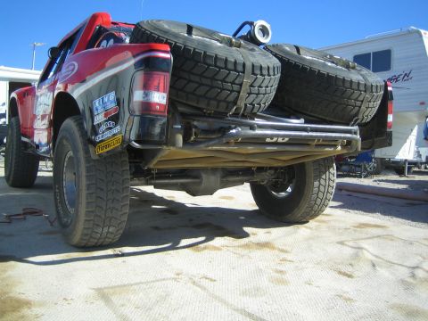 1990 Ford Bronco - Rear view of a lifted off-road race truck exposing its underside, dual spare tires and rear suspension in a pit area.