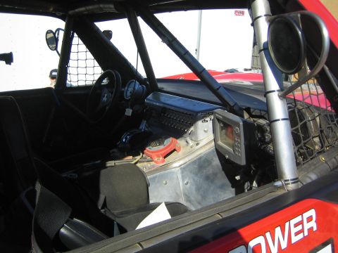 1990 Ford Bronco - Cockpit of an off-road race truck showing the steering wheel, mounted GPS screen, switch panel and roll cage netting.