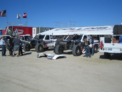 1990 Ford Bronco - Several off-road race trucks parked under a Collins canopy in a desert staging area with American and other flags flying.