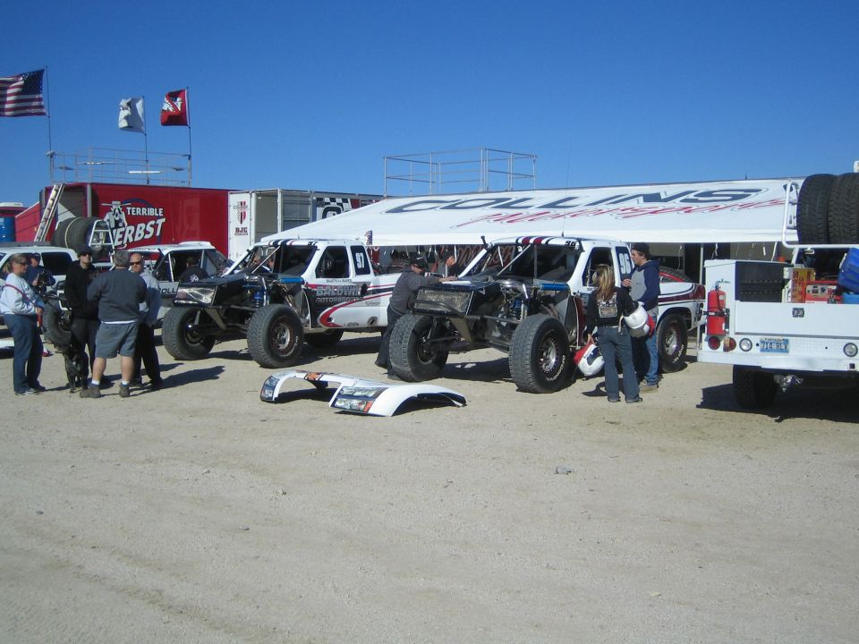 Row of off-road race trucks under a canopy in a desert pit with flags
