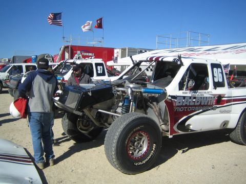 1990 Ford Bronco - white number 96 Baldwin Motorsports off-road race truck with its rear engine and suspension exposed in a desert pit.