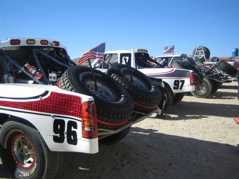 1990 Ford Bronco - White off-road race trucks numbered 96 and 97 lined up rear-first in a desert pit, each carrying spare tires and US flags.