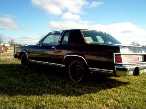 1985 Ford LTD Crown Victoria - The rear of a dark-colored sedan is shown parked on a grassy area under a partly cloudy sky.