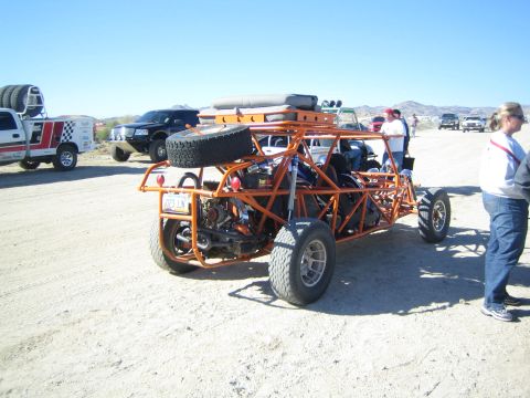 1990 Ford Bronco - orange tube-frame off-road buggy with roof-mounted spare tires and lights parked on a desert flat with people nearby.