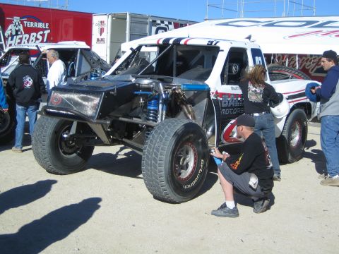 1990 Ford Bronco - black-and-white open-wheel off-road race truck sits in a pit as a kneeling man in black works beside it near trailers.