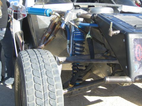1990 Ford Bronco - Close-up of a race truck's front suspension showing a blue coilover, bypass shocks, A-arms and a wide off-road tire.