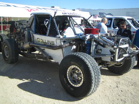 1990 Ford Bronco - white Collins open-wheel off-road race truck with exposed rear engine and tube chassis parked in a desert pit.