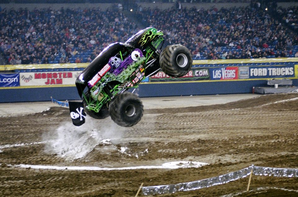 Monster truck with skull graphics airborne over dirt track, crowd in background