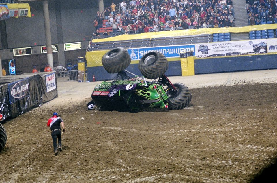 Overturned green monster truck with flame graphics on dirt track, audience in background