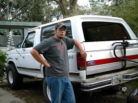1986 Ford Bronco - A man leans against a white Ford Bronco with a red stripe and a bike rack on the back.