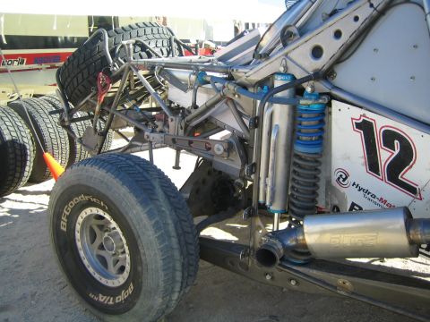 1990 Ford Bronco - Rear suspension and exhaust of a silver tube-chassis race truck marked 12, with spare tires stacked alongside in a pit.