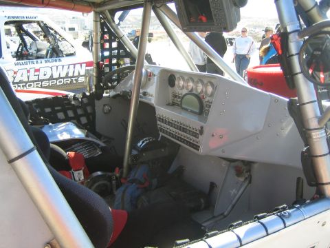 1990 Ford Bronco - Cockpit of a Baldwin Motorsports race truck showing a digital gauge panel, switches and seat as people stand outside.