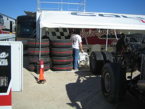 1990 Ford Bronco - Stacks of off-road race tires sit under a Collins Motorsports canopy beside a trailer as a man stands nearby.