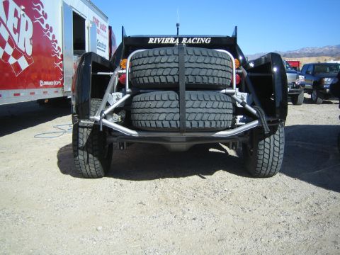 1990 Ford Bronco - Rear view of a black off-road race truck labeled Riviera Racing carrying two spare tires on a tube rack in a desert pit.