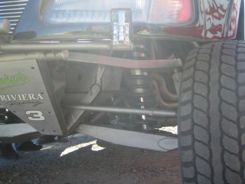 1990 Ford Bronco - Rear suspension of a Riviera Racing number 3 off-road truck showing leaf spring, coilover and tube linkages behind a large knobby tire.