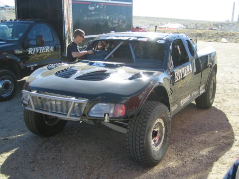 1990 Ford Bronco - Black Riviera Racing off-road trophy truck with vented hood and tube grille parked beside a team trailer at a dusty desert event.