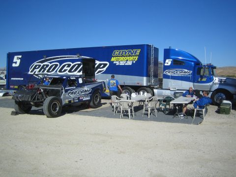 1990 Ford Bronco - Blue and silver Pro Comp number 5 off-road race truck parked near a Coyne Motorsports semi-truck and trailer with crew seated at tables.