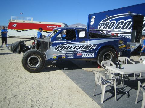 1990 Ford Bronco - Blue and silver Pro Comp number 5 off-road race truck on a salt-flat paddock beside its team trailer with crew in blue shirts nearby.