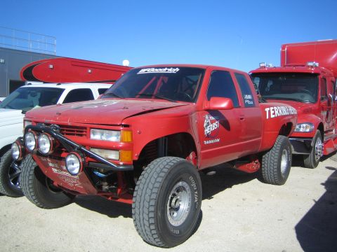 1990 Ford Bronco - Red lifted Chevrolet extended-cab off-road race truck with a roof light bar and Herbst decals parked beside trailers in a desert lot.