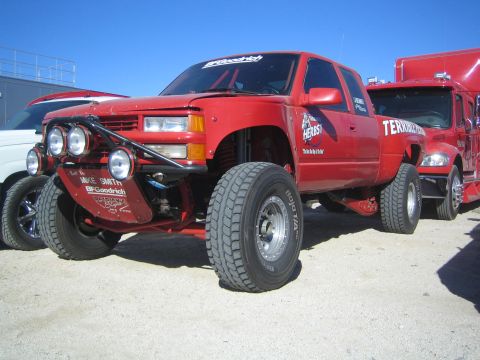 1990 Ford Bronco - Front three-quarter view of a red lifted Chevrolet off-road race truck with auxiliary lights and tall tires parked at a desert race event.