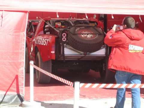 1990 Ford Bronco - Rear view of a red Terrible Herbst off-road race truck carrying a spare tire, photographed by a man in a red Herbst jacket under a tent.